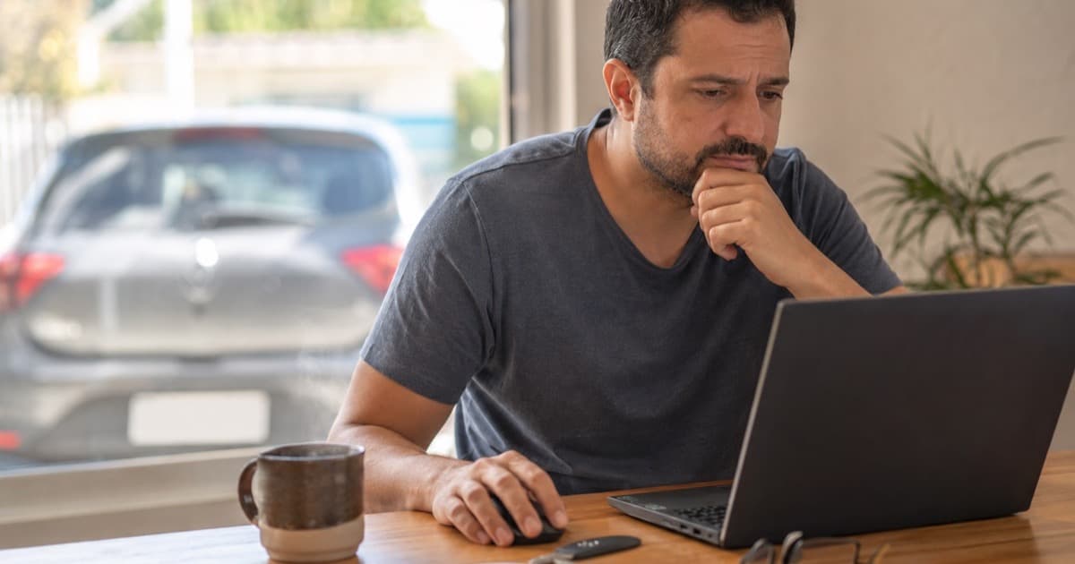 Homem pesquisa no notebook em casa se é crime comprar a placa do carro pela internet, com a chave do veículo sobre a mesa e um carro desfocado ao fundo
